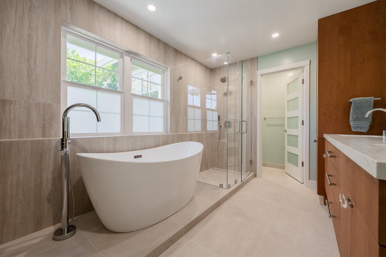 After: Remodeled primary bathroom by ASL Remodeling featuring freestanding soaking tub, frameless glass shower, travertine-look tile walls, and wood vanity with quartz top