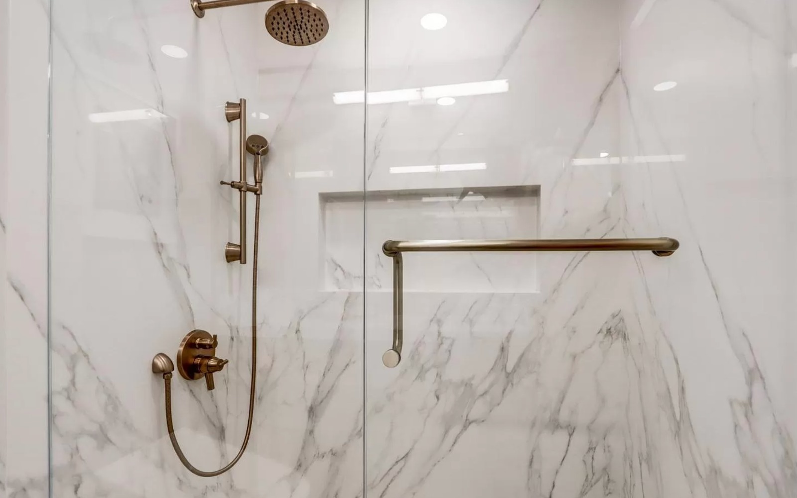 Experienced remodeling bathroom contractors created spa-inspired walk-in shower with floor-to-ceiling marble tile, elegant brass grab bars, built-in niches, and handheld shower.