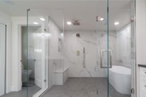 Sleek bath and shower remodel embraces minimalist aesthetic with floor-to-ceiling frameless glass enclosure, elegant freestanding tub, and white porcelain slabs.