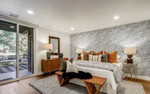 Spacious master bedroom addition featuring dramatic stone accent wall, contemporary furnishings, recessed lighting, and sliding glass doors opening to outdoor patio.