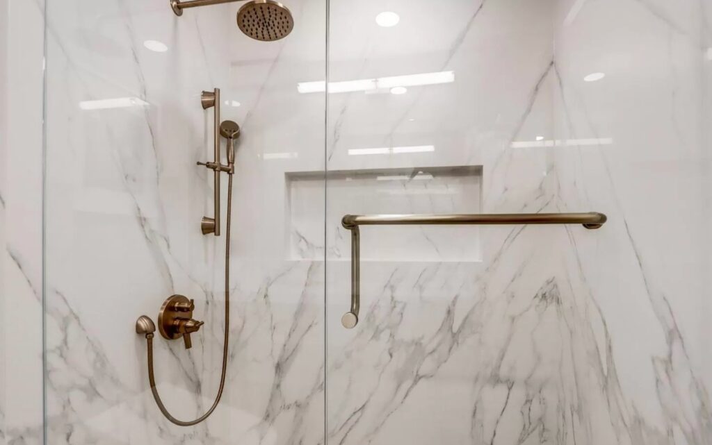 Experienced remodeling bathroom contractors created spa-inspired walk-in shower with floor-to-ceiling marble tile, elegant brass grab bars, built-in niches, and handheld shower.