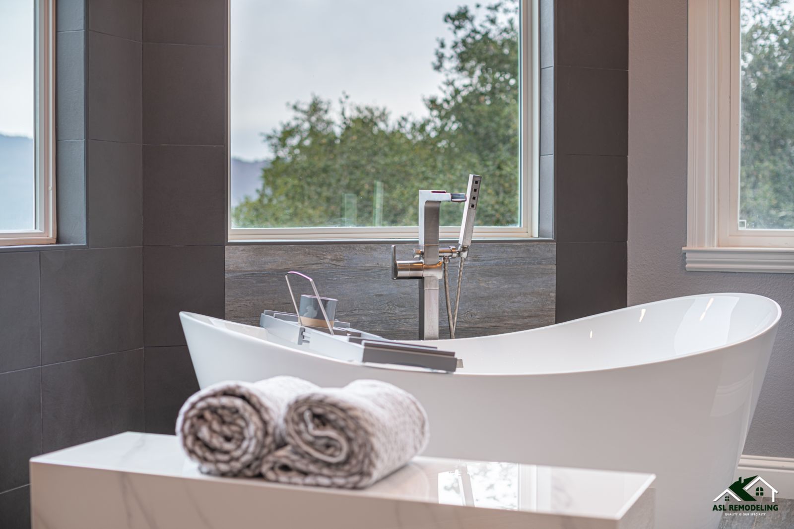 Freestanding bathtub with modern chrome faucet near large windows, with rolled towels in the foreground.