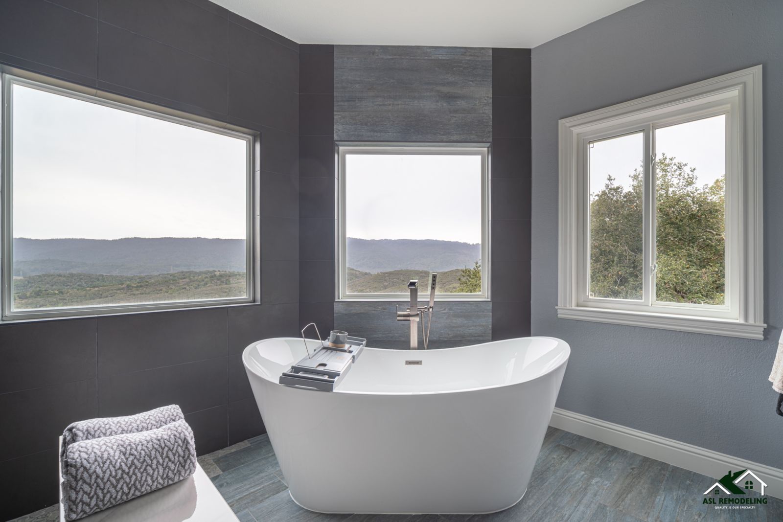 Modern bathroom with freestanding tub centered between three large windows overlooking scenic mountain and forest views.