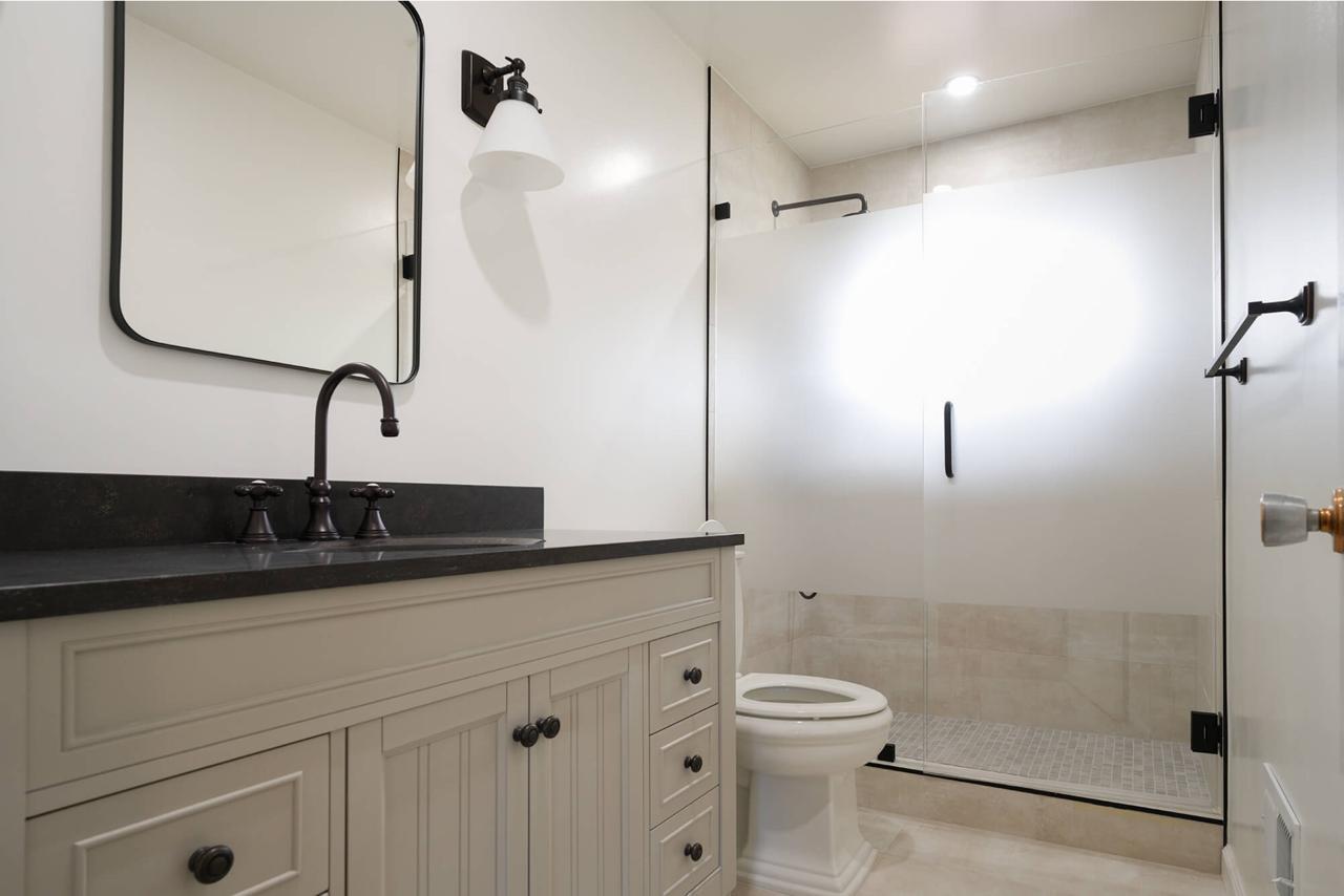 Modern guest house bathroom with permit coordination for plumbing upgrades featuring white shaker cabinets, black countertop, frameless shower, and contemporary plumbing fixtures with black hardware accents.