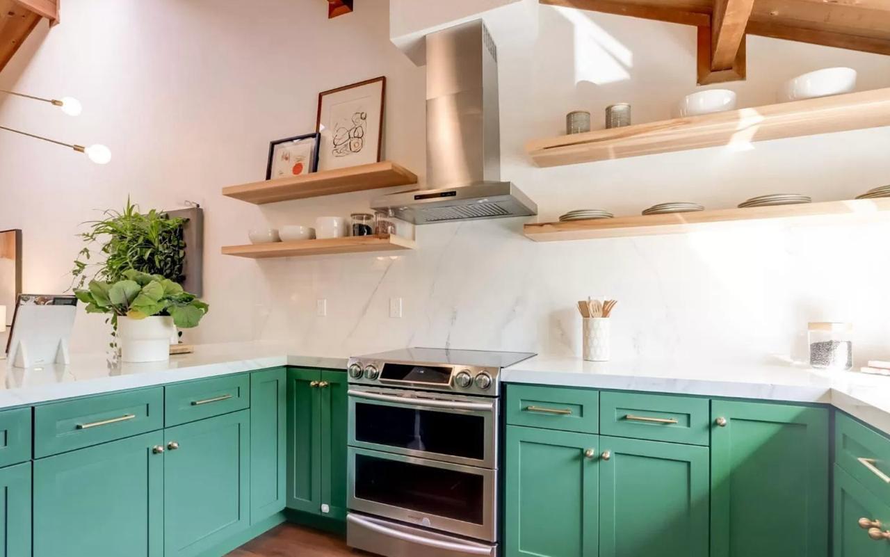 Rental unit modern kitchen featuring bold green lower cabinets, white upper cabinets, open wood shelving, porcelain slabs backsplash, and stainless appliances.